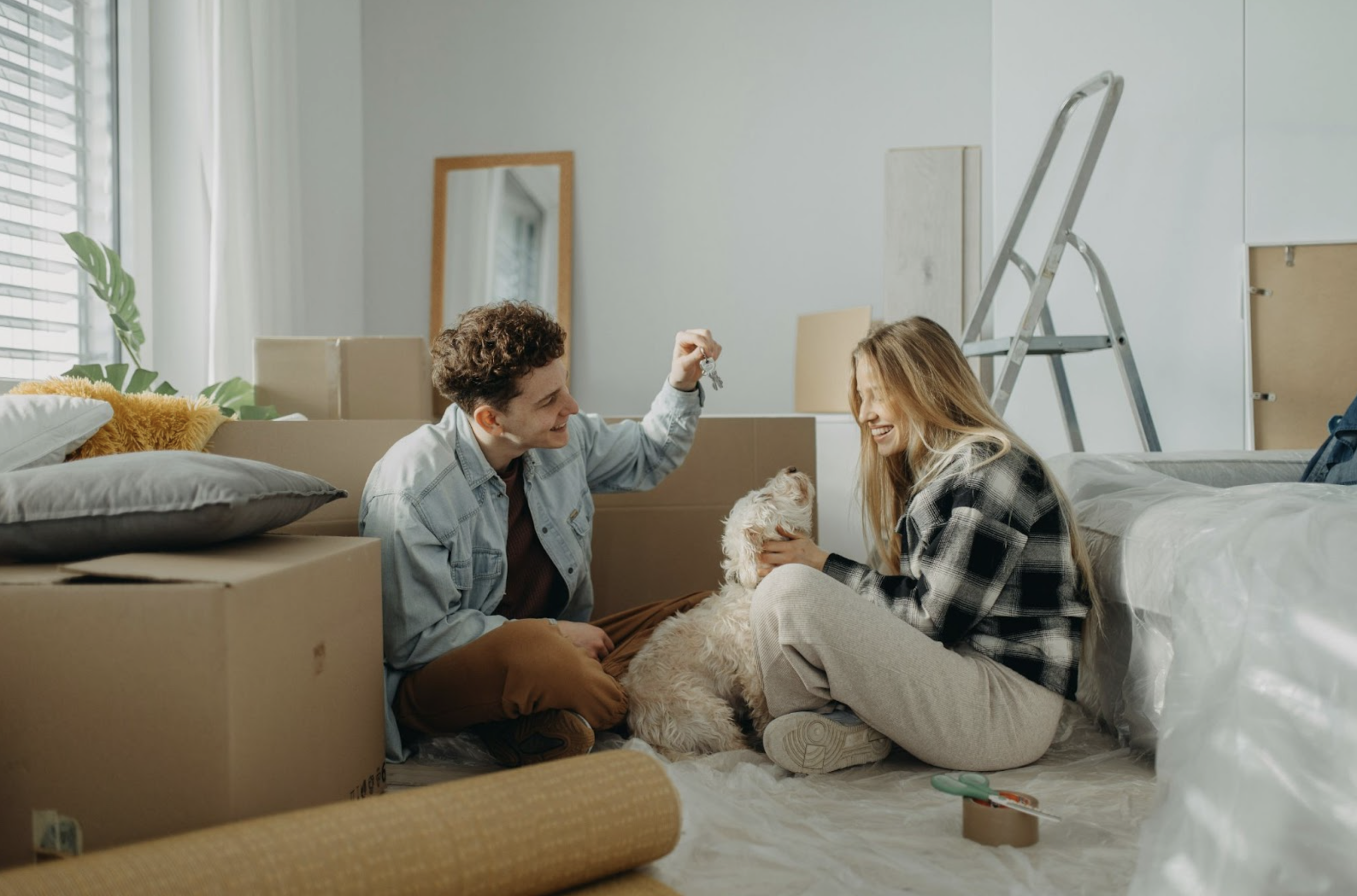 A young couple sitting on the floor surrounded by moving boxes, smiling and holding up a house key, with a small dog beside them.